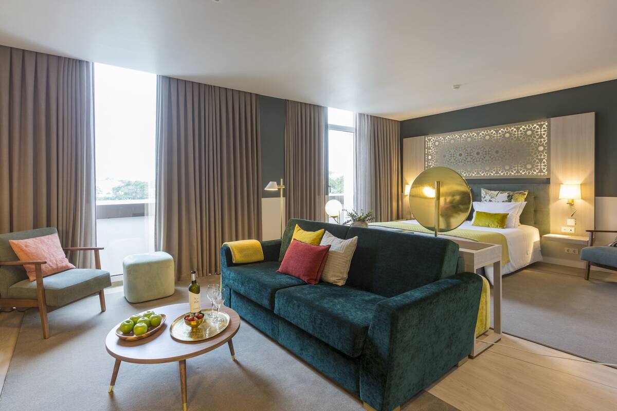 A modern hotel room with a green sofa, a dining table with fruit and wine, a bed with a headboard, and a view of the outdoors.