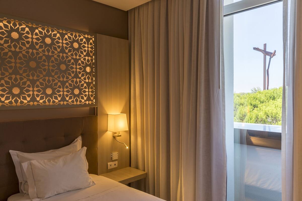 A hotel room with a bed, a nightstand with a lamp, a decorative wall panel, and a view of greenery and a cross outside the window.