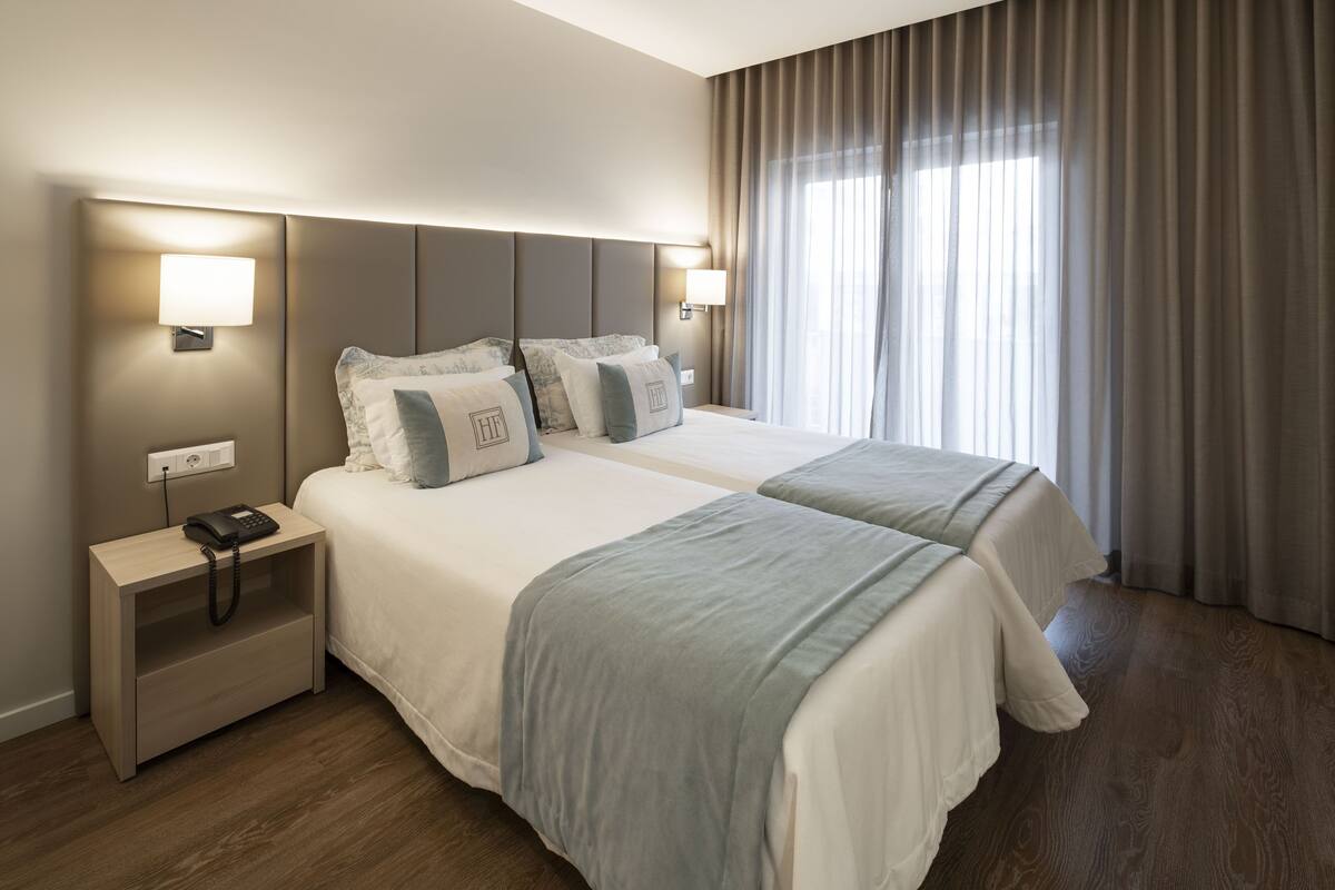 Quarto de hotel com duas camas, luminárias de cabeceira, mesa de cabeceira com telefone e janela com cortinas.