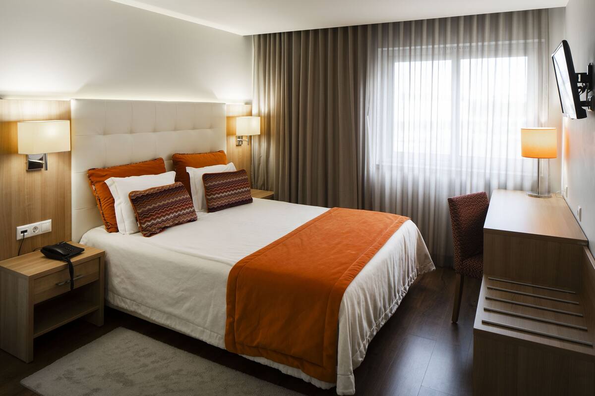 Quarto de hotel com uma cama grande, duas mesas de cabeceira com luminárias, uma escrivaninha com cadeira e uma janela ampla com cortinas.