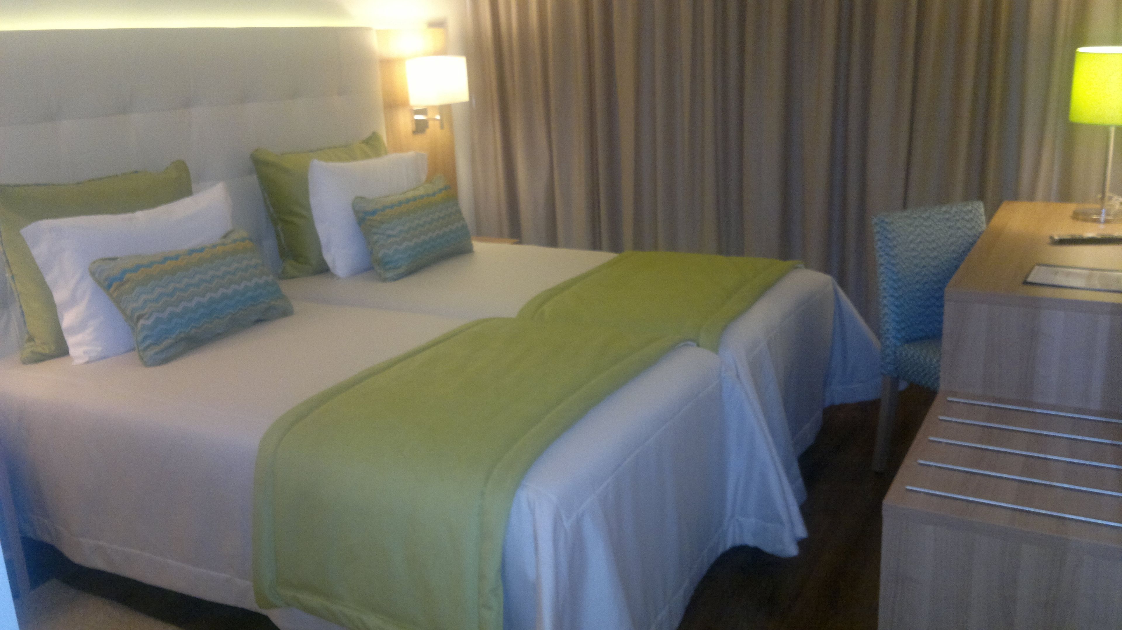 Quarto de hotel com duas camas, cada uma com roupa de cama verde e branca, uma escrivaninha de madeira e uma luminária.