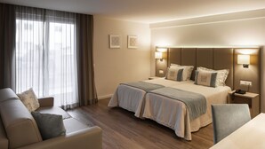 Habitación de hotel con cama, dos mesitas de noche, un escritorio, una silla, un sofá y una ventana con cortinas.