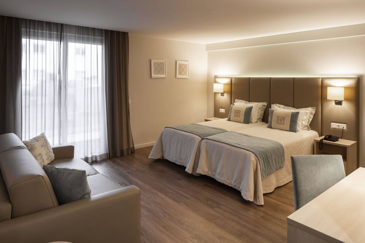 Quarto de hotel com cama, duas mesas de cabeceira, escrivaninha, poltrona, sofá e janela com cortinas.