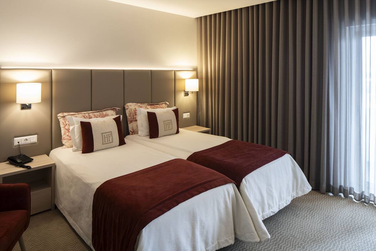 Quarto de hotel com duas camas, uma mesa de cabeceira com telefone e vista para o exterior através de cortinas.