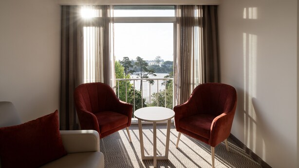 Habitación de hotel con balcón, dos sillones rojos, una mesita redonda y vistas a un paisaje urbano.