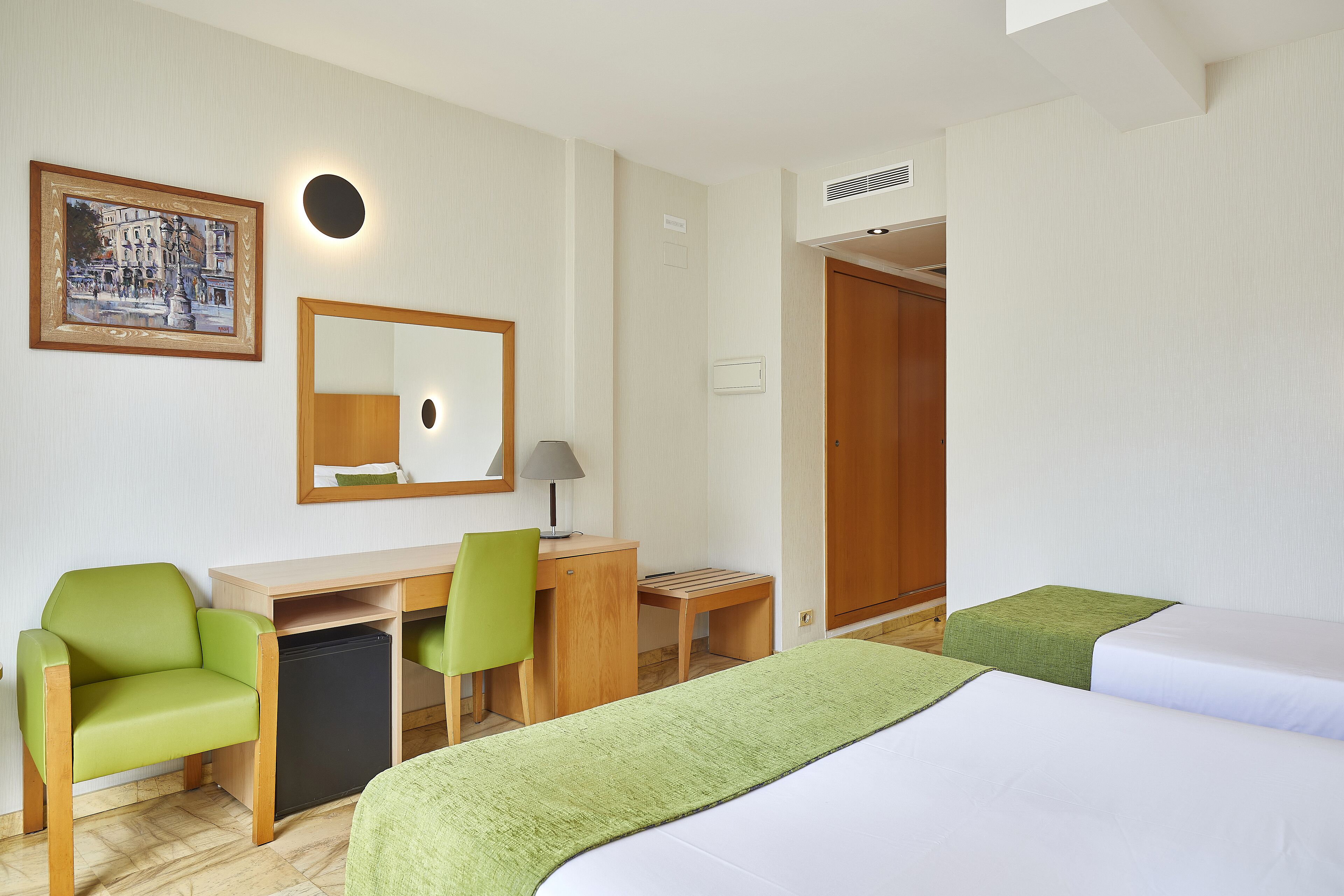 A hotel room with a desk, two green chairs, a bed, and a painting on the wall.