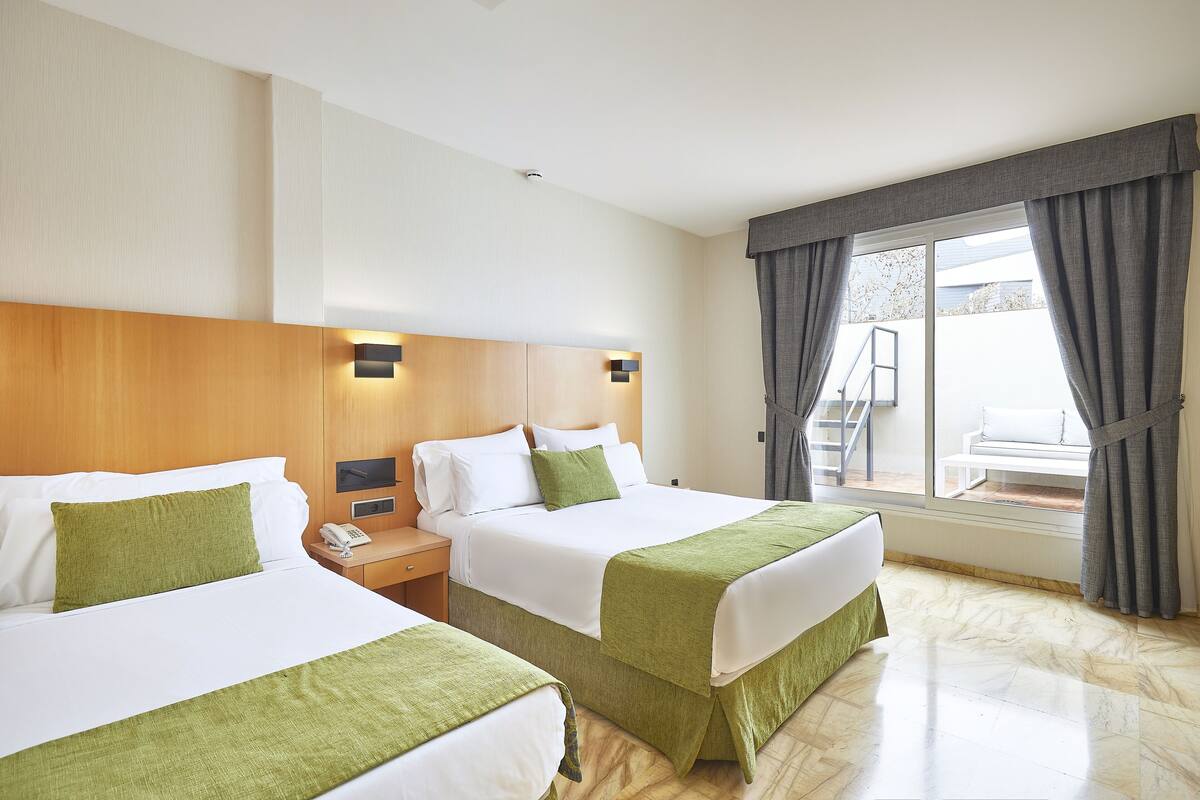 A hotel room with two beds, a wooden headboard, a nightstand with a phone, and a view of the outdoors through a large window with curtains.