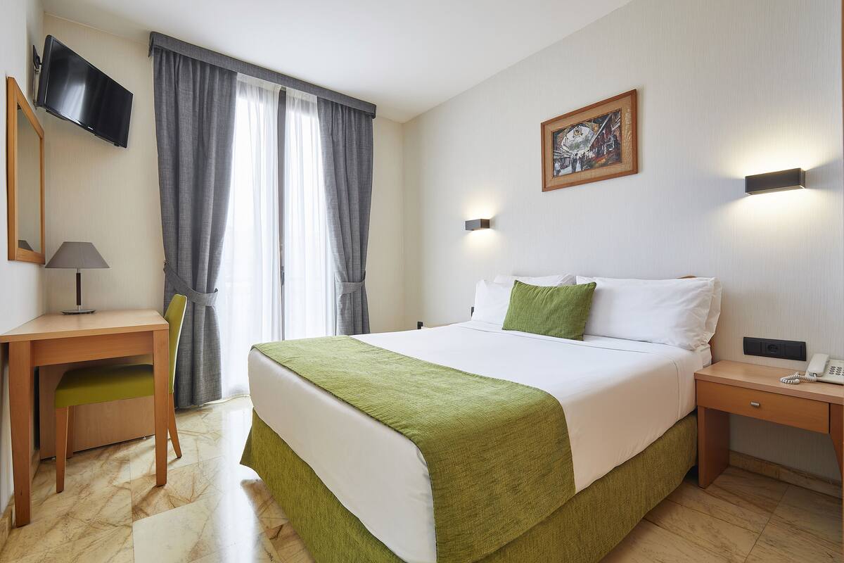 A hotel room with a bed, a desk, a chair, a TV, a painting, and a phone on the nightstand.