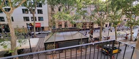 A street view with a building labeled "LA QUILLA DE OFFICE" in the background, a metal fence in the foreground, and trees lining the street.
