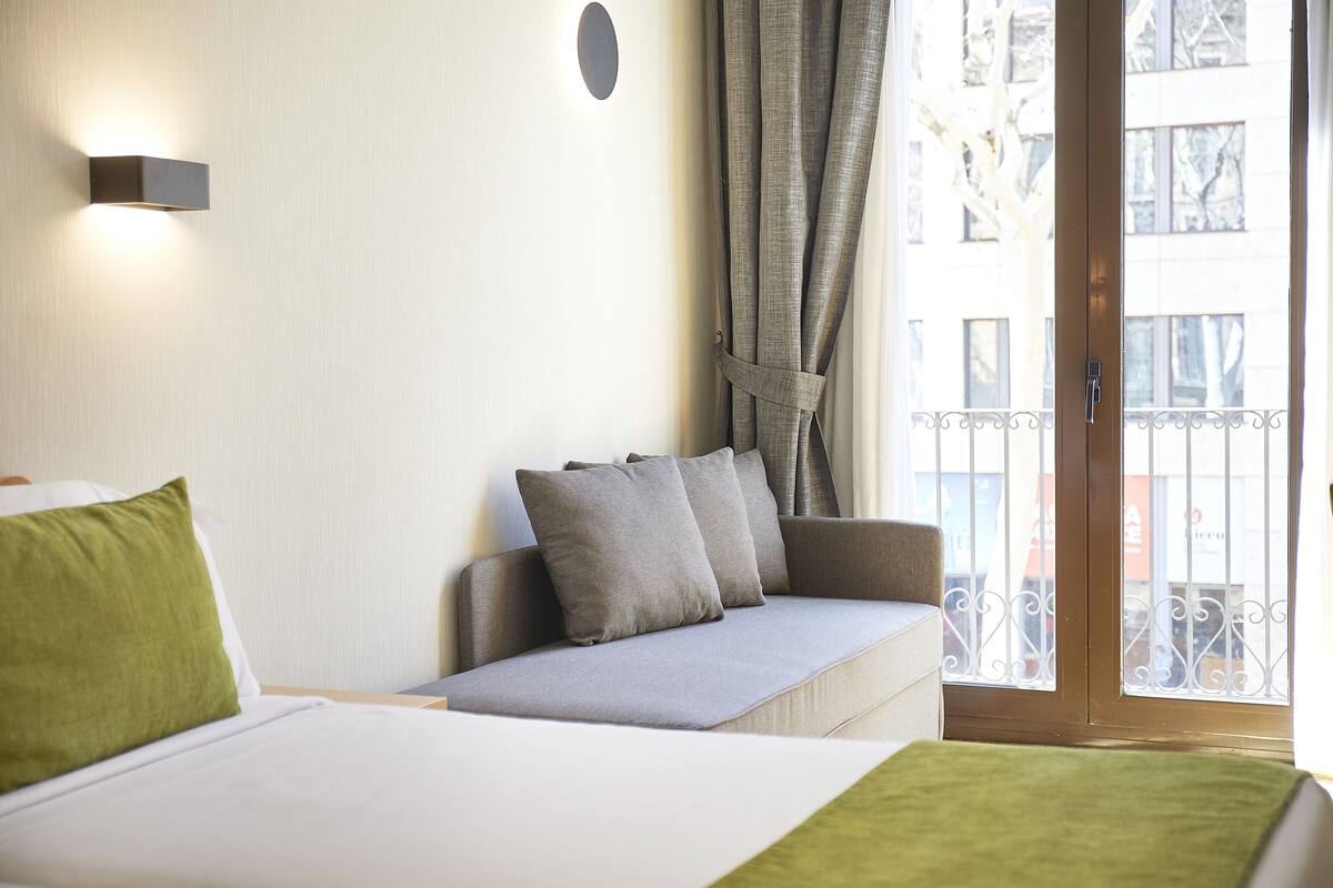 A bedroom with a bed, a sofa, a window with curtains, and a balcony.
