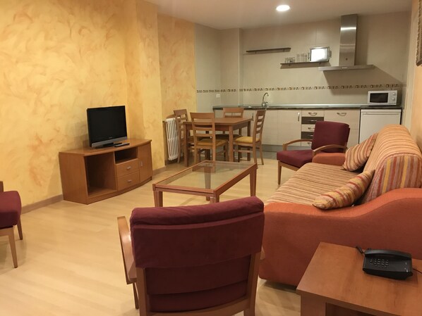A living room with a sofa, chairs, a coffee table, a TV, and a kitchen area with a microwave and stove.