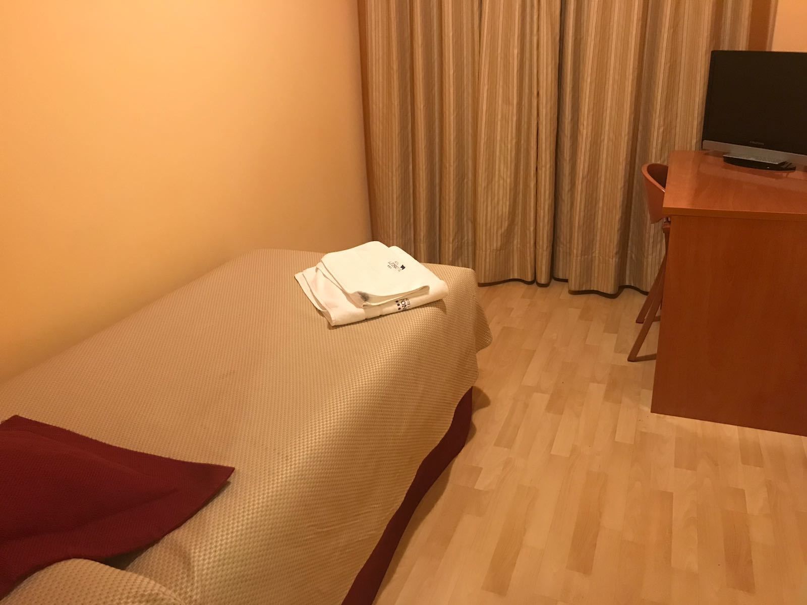 A small, neatly made bed with a white blanket and a red pillow, a wooden desk with a television, and a window with curtains.