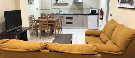A living room with a sofa, a TV, a dining table with chairs, and a kitchen area with cabinets and a microwave.