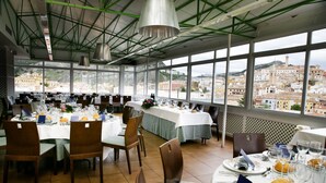 Un restaurant amb diverses taules preparades per dinar, grans finestres que ofereixen vista a un paisatge urbà i un sostre alt amb bigues al descobert.
