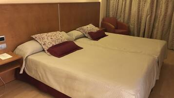 Una cama doble con una colcha color beige y dos almohadas, una mesita de noche de madera con una lámpara, y una silla roja en una habitación de hotel.