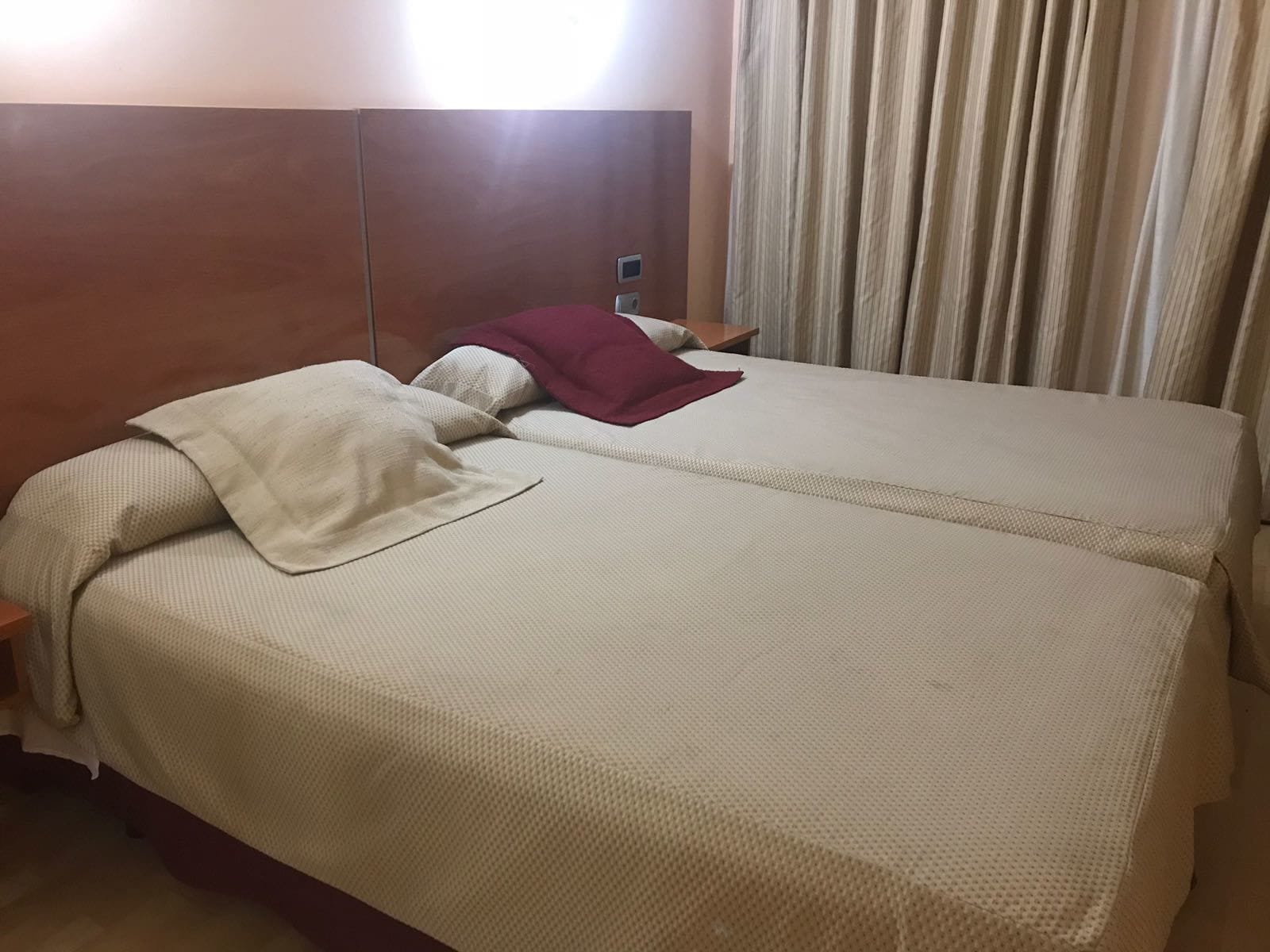 A double bed with a beige bedspread and a red pillow, a wooden headboard, a nightstand, and a window with curtains.