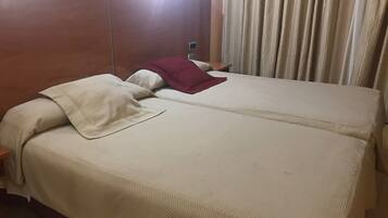 A double bed with a beige bedspread and a red pillow, a wooden headboard, a nightstand, and a window with curtains.