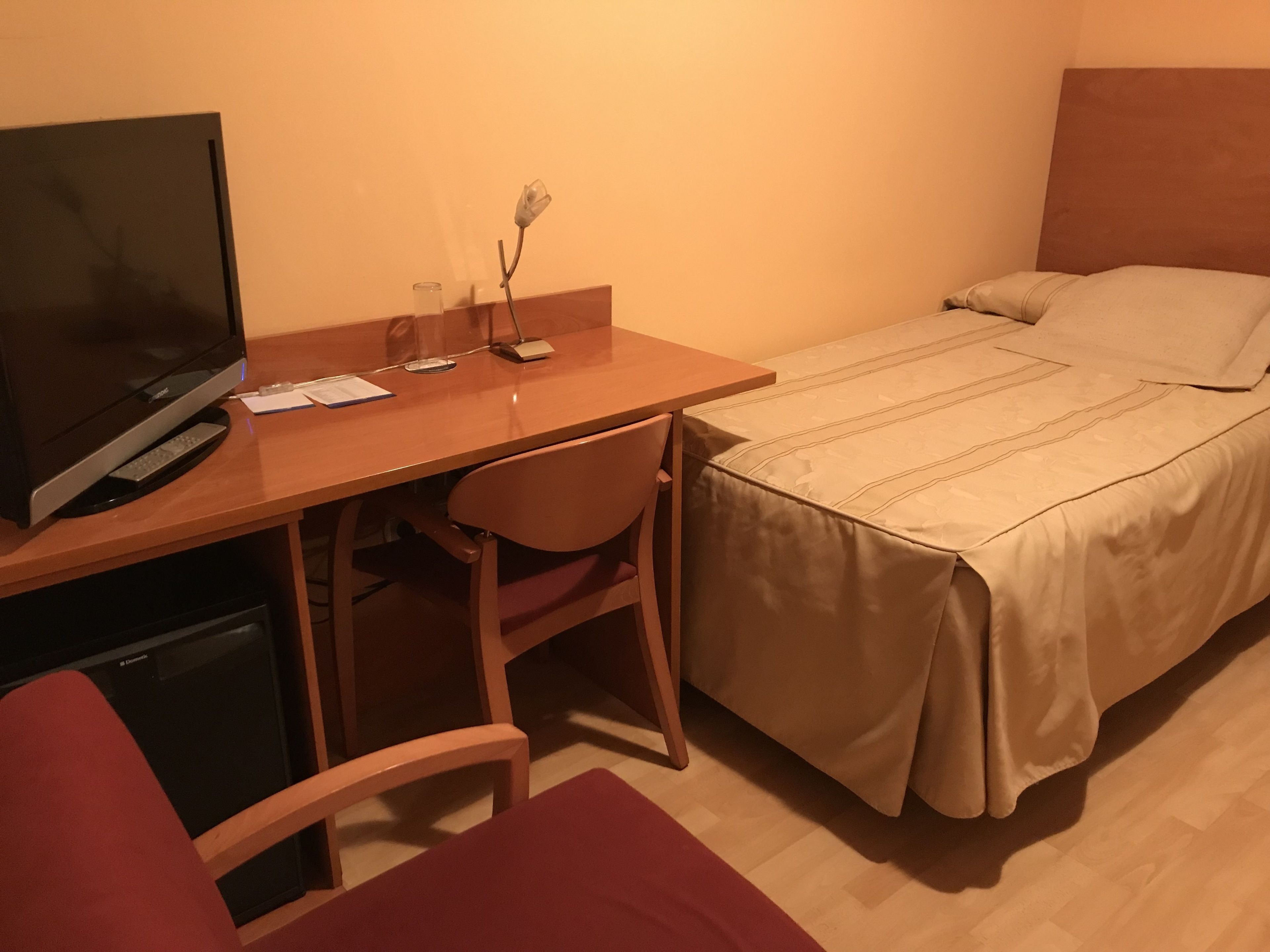 Ein Hotelzimmer mit Holzschreibtisch, Fernseher, einem Bett mit beige farbenem Überwurf und einer kleinen Vase mit einer Blume.