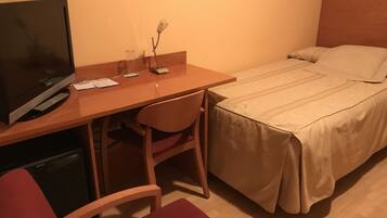 Ein Hotelzimmer mit Holzschreibtisch, Fernseher, einem Bett mit beige farbenem Überwurf und einer kleinen Vase mit einer Blume.