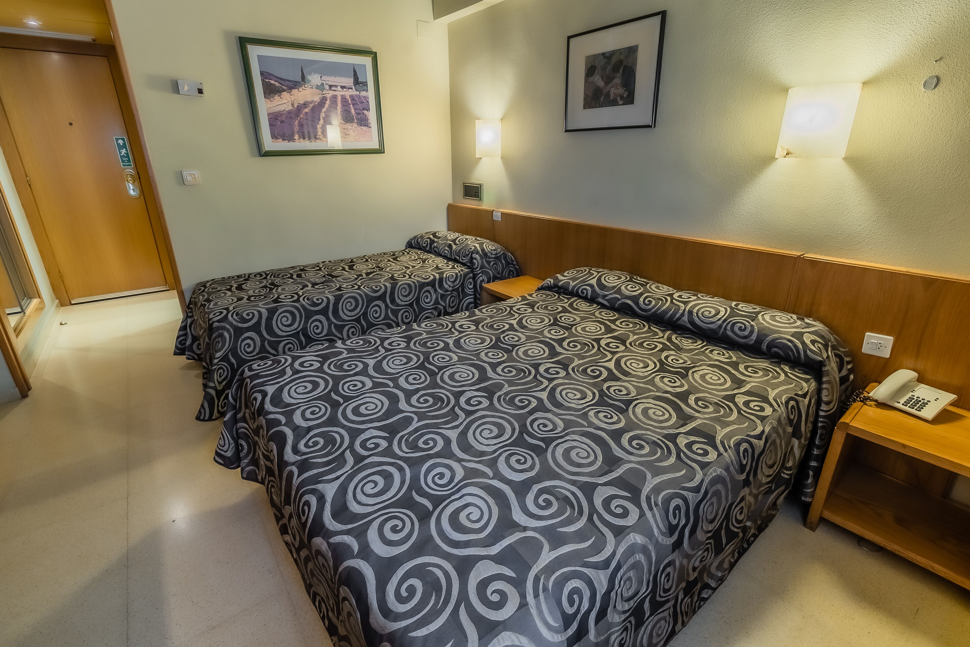 Hotelkamer met twee bedden, een houten hoofdbord, een nachtkastje met telefoon en twee wandlampen.