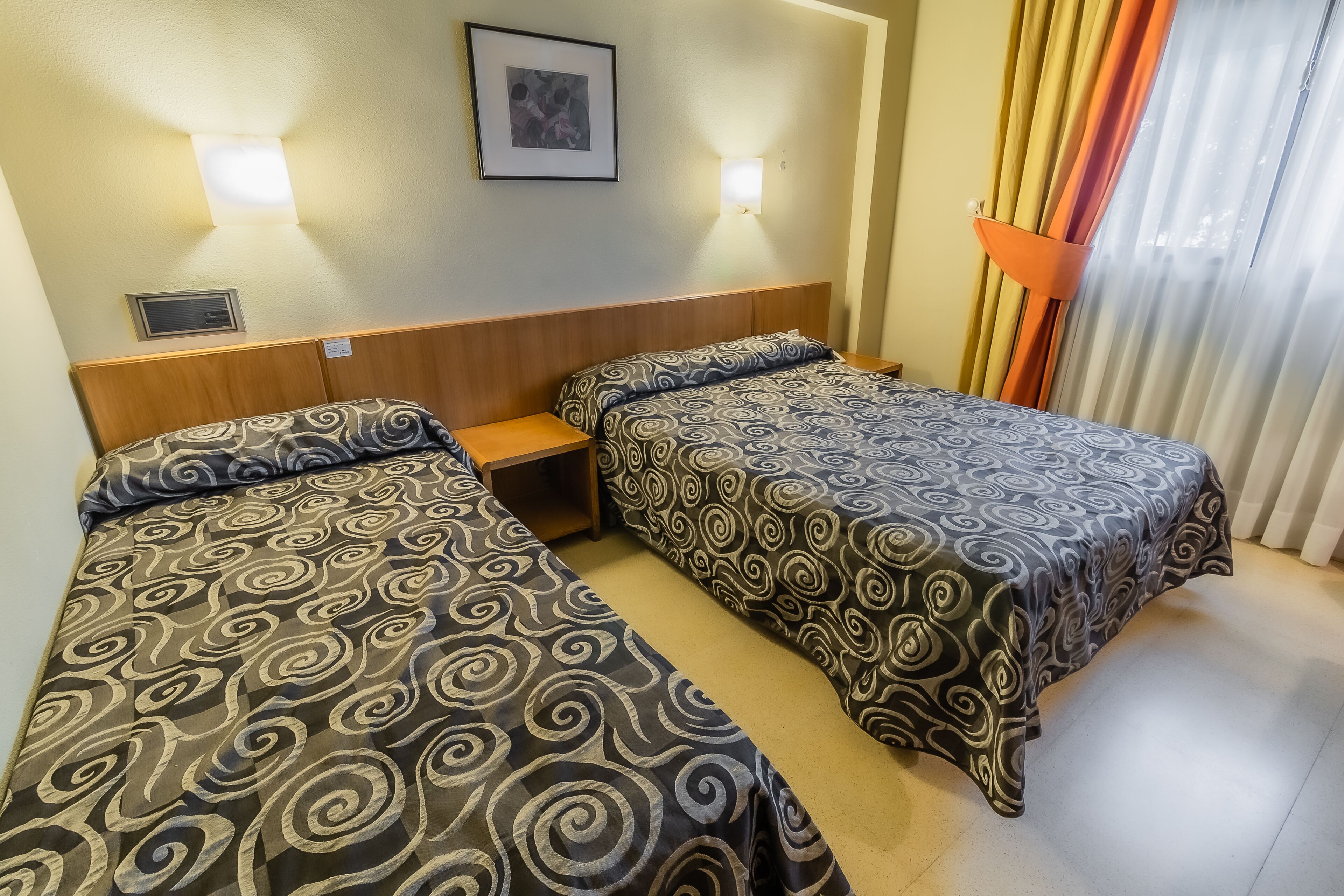 A hotel room with two beds, each with patterned bedding, a headboard, a bedside table, a wall-mounted light, and a window with curtains.