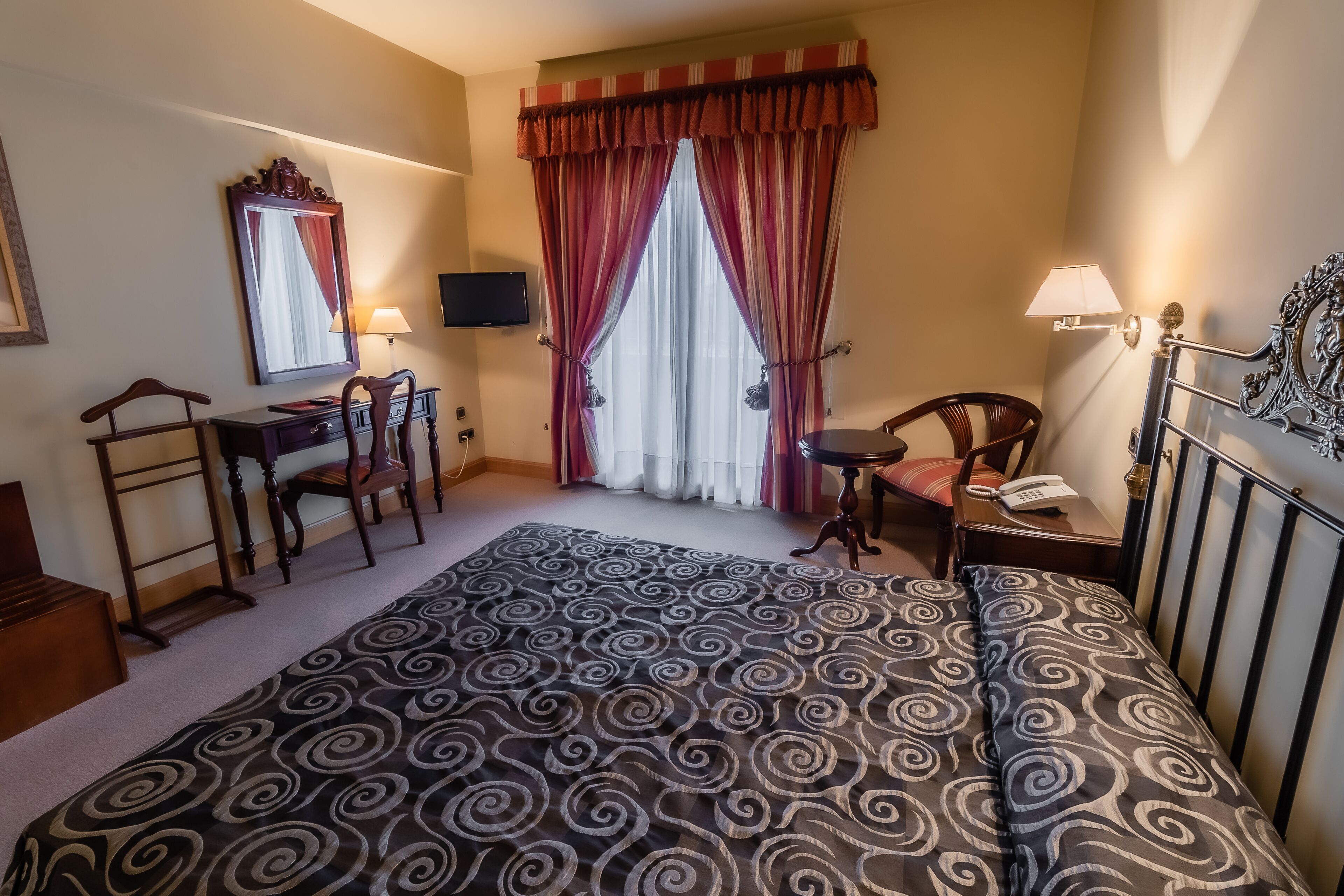 A hotel room with a large bed, a desk with a chair, a TV mounted on the wall, and a window with curtains.