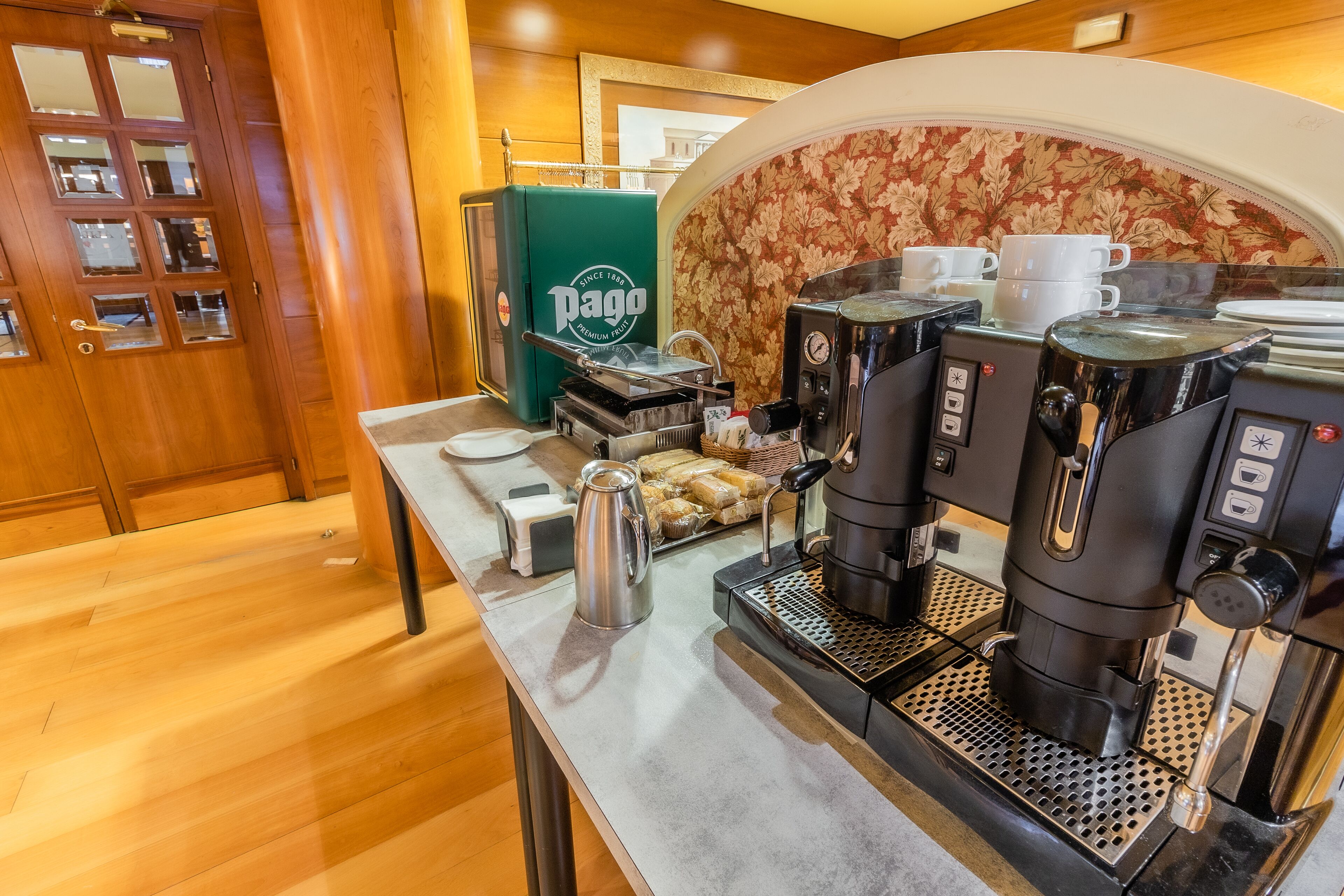 Uma estação de café com várias máquinas de espresso, uma máquina de café verde da marca 'Pago', e uma bandeja de pastelaria.