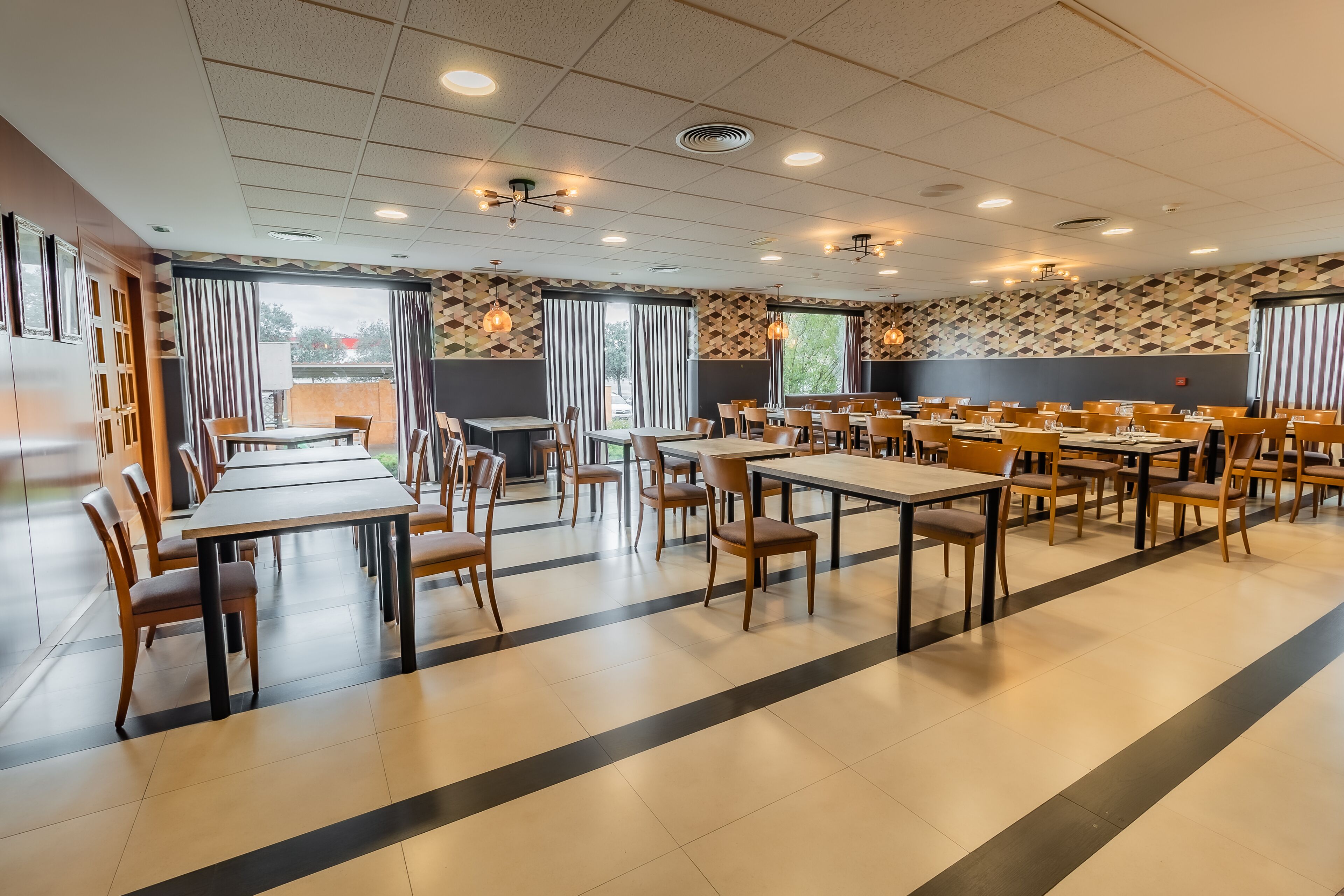 A spacious dining area with multiple tables and chairs, large windows with curtains, and a patterned wall design.