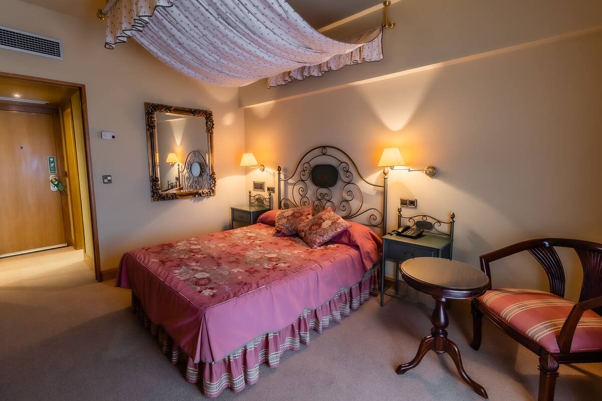 A bedroom with a bed, two bedside tables with lamps, a chair, a mirror, and a door.