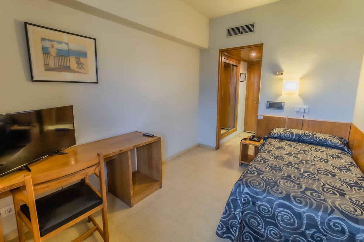 A hotel room with a bed, a desk, a TV, a wall-mounted lamp, and a framed picture.