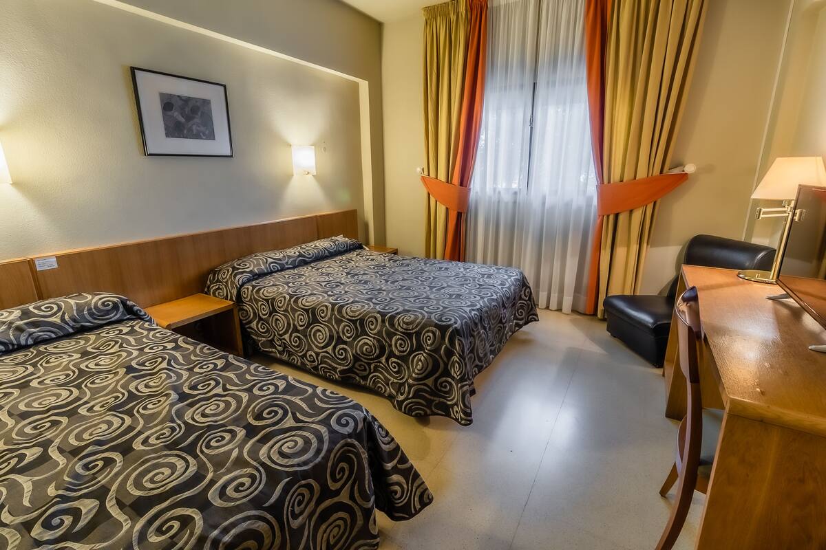 A hotel room with two beds, a desk, a chair, and a lamp.