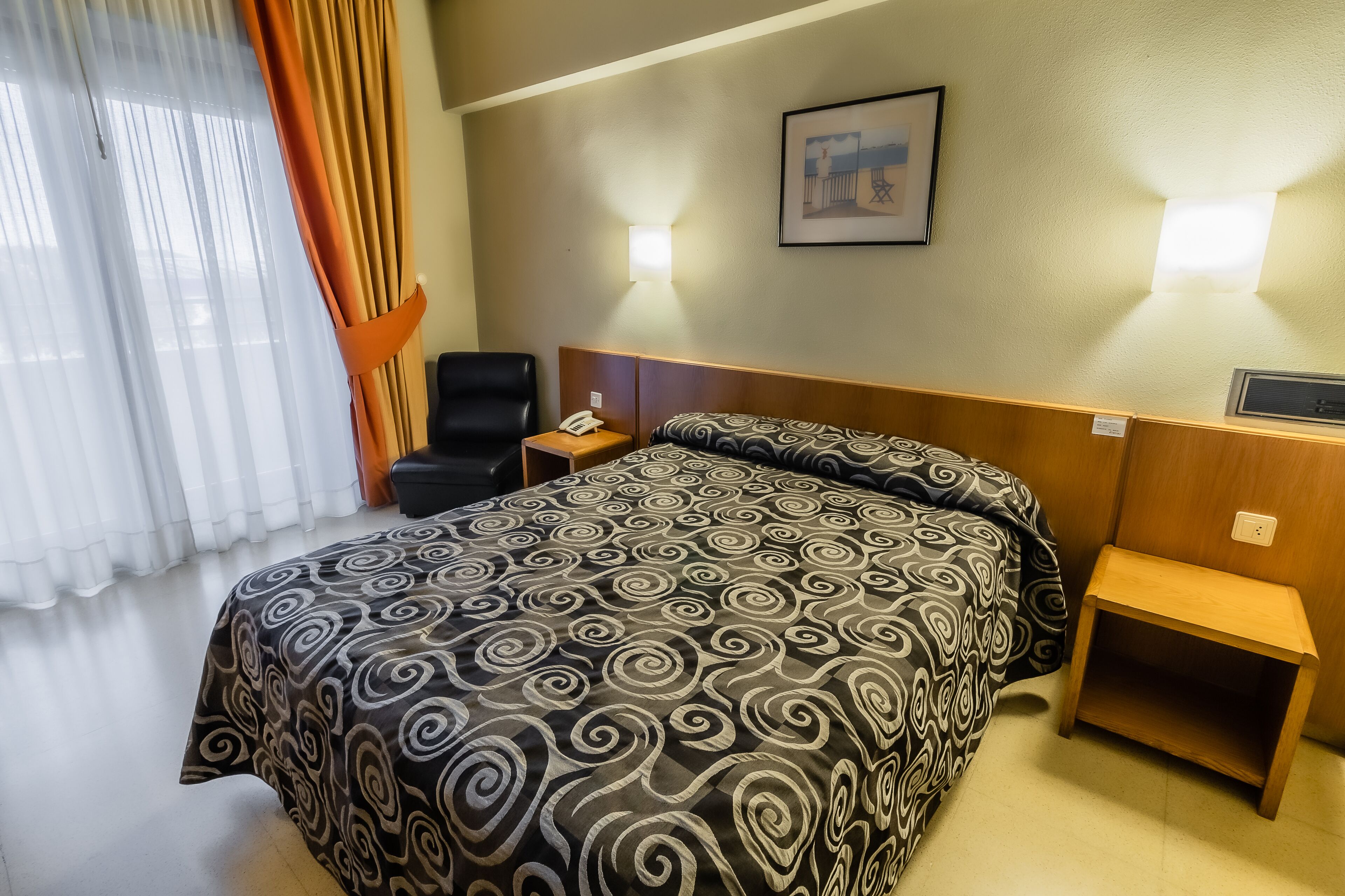 Ein Hotelzimmer mit einem großen Bett, einem Nachttisch mit Telefon, einem Sessel und einem Bild an der Wand.