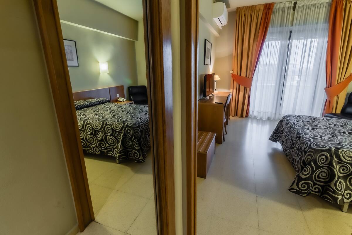 A hotel room with two beds, a desk, a chair, a lamp, and a window with curtains.