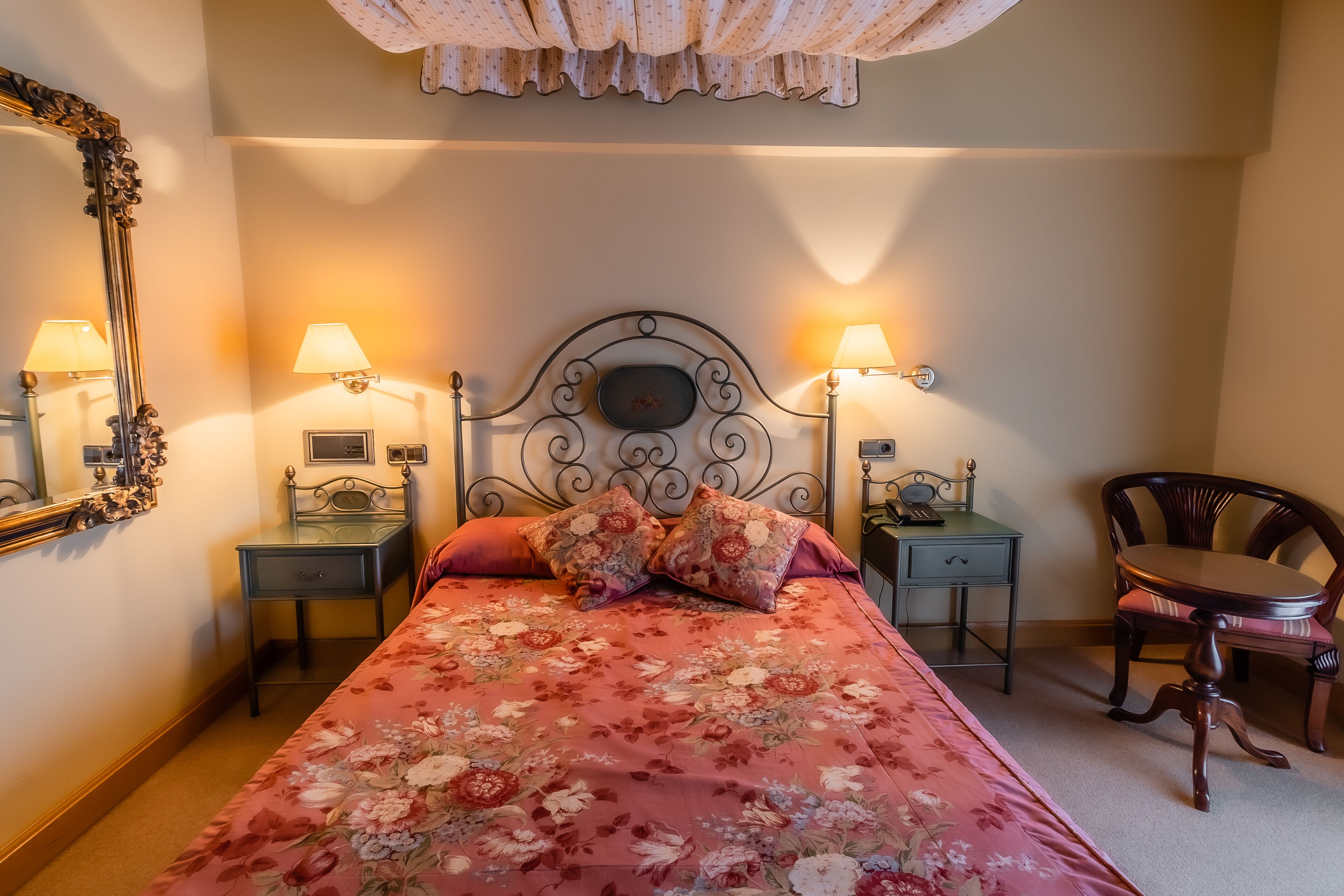 A neatly arranged bedroom with a floral bedspread, two bedside lamps, a mirror, a small table, and a chair.