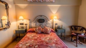 A neatly arranged bedroom with a floral bedspread, two bedside lamps, a mirror, a small table, and a chair.