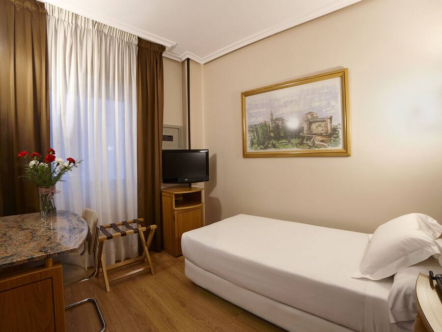 Habitación de hotel con cama, televisión, un cuadro, escritorio con silla y un jarrón de flores.