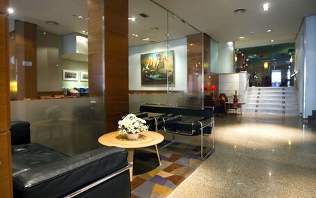 A modern lobby with a glass partition, a seating area with a round table, and a staircase leading to an upper floor.