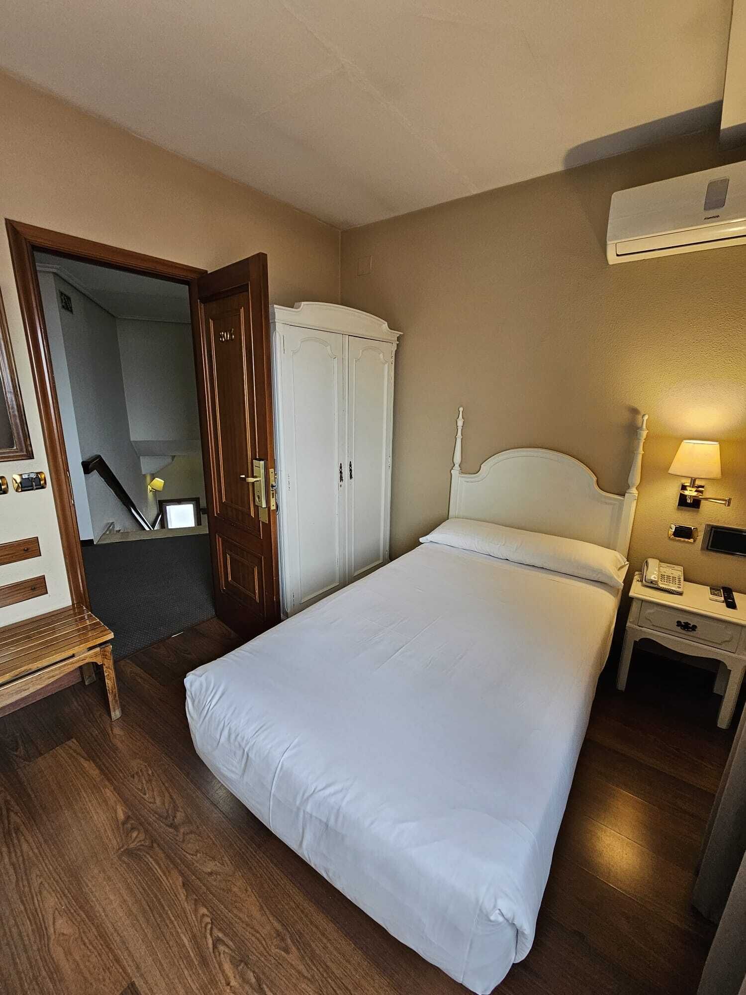 A hotel room with a bed, a wardrobe, a bedside table with a lamp, and a door leading to a staircase.