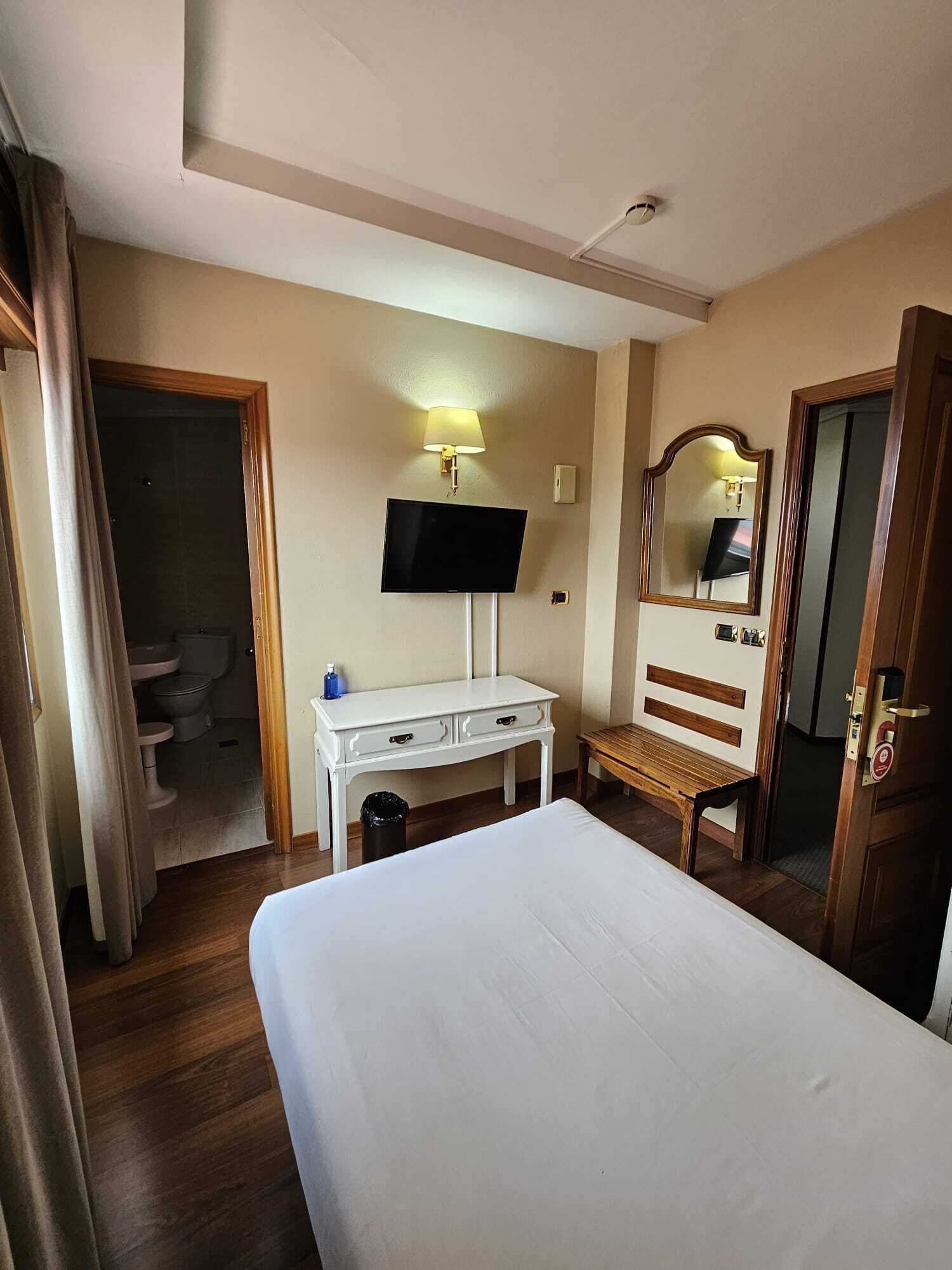 Hotelkamer met een bed, een bureau met een flatscreen televisie, een bankje, een spiegel en een deur.