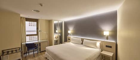 A hotel room with a large bed, a desk, a chair, and a window with curtains.
