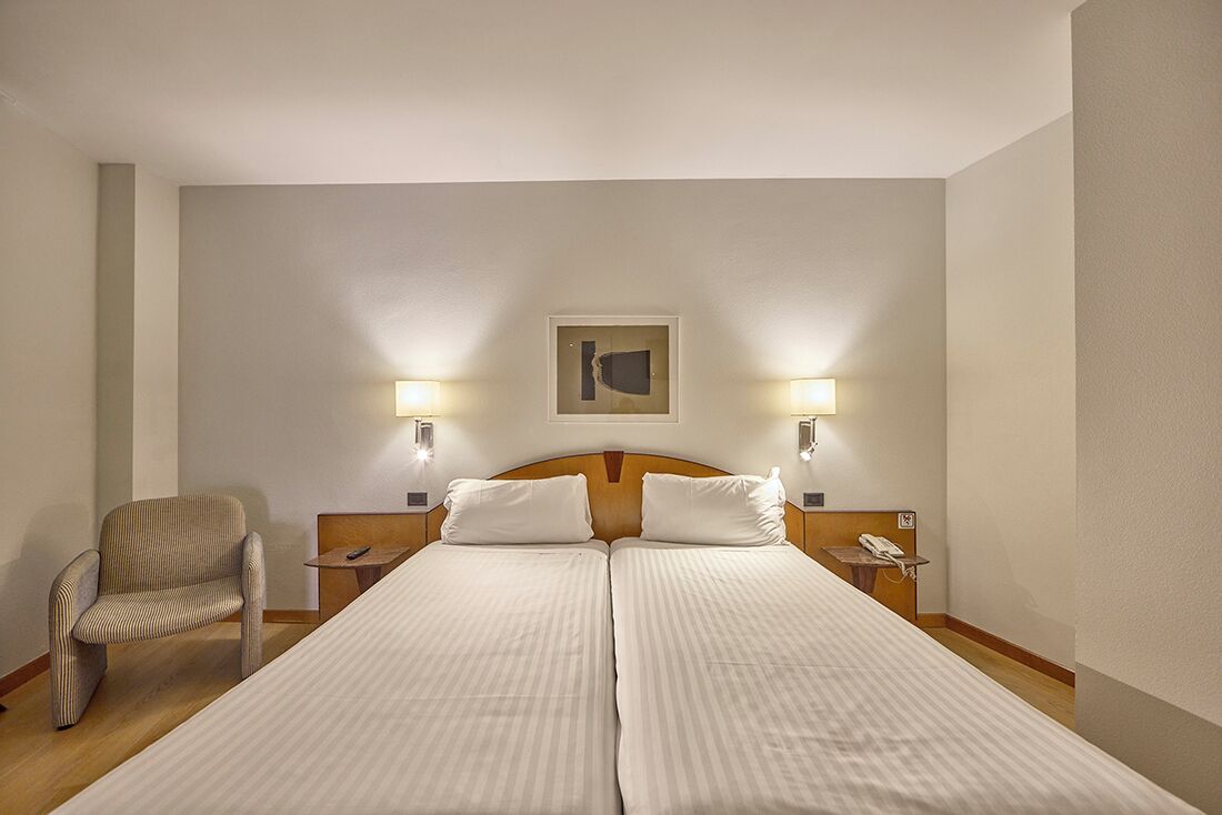 Un lit double avec deux oreillers, une chaise, des tables de chevet avec des lampes et une œuvre d’art accrochée au mur.
