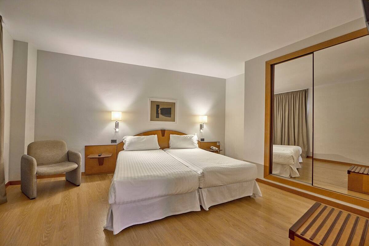 A hotel room with a double bed, a chair, a nightstand, a mirror, and a wooden floor.