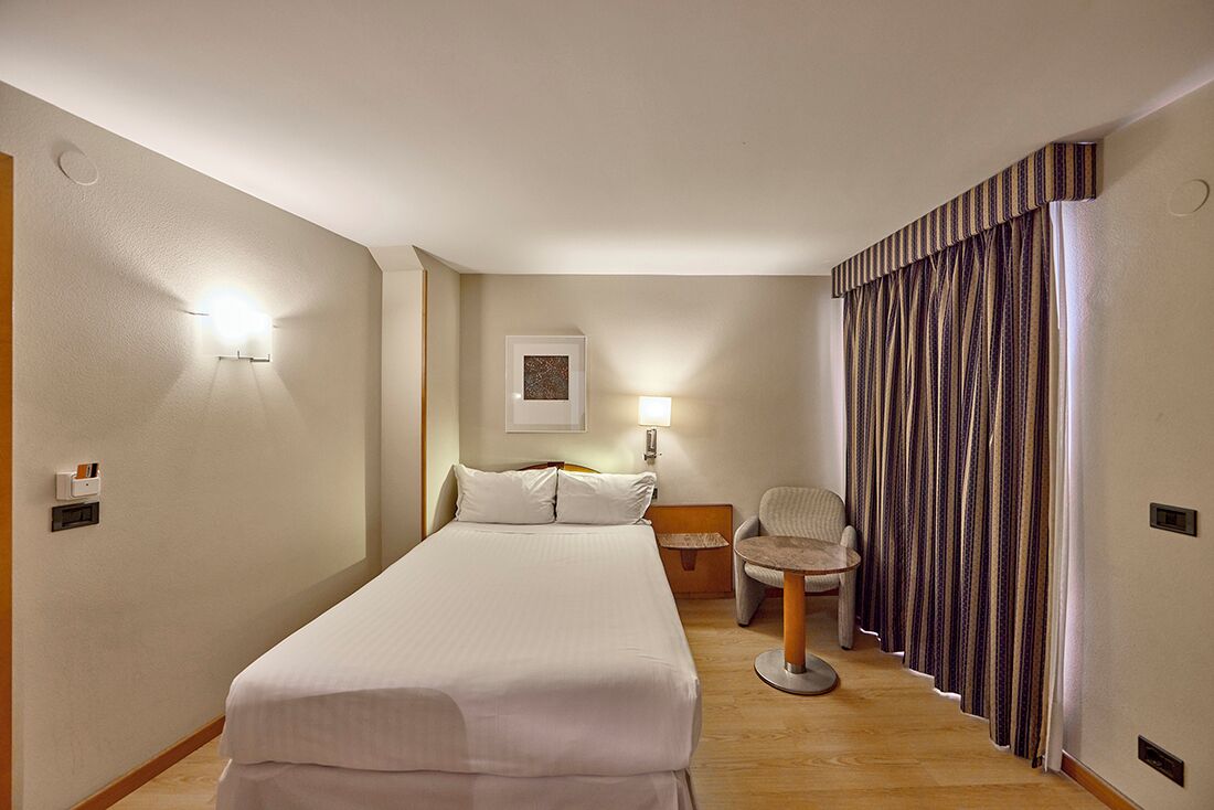 A hotel room with a large bed, a chair, a desk, and a window with curtains.