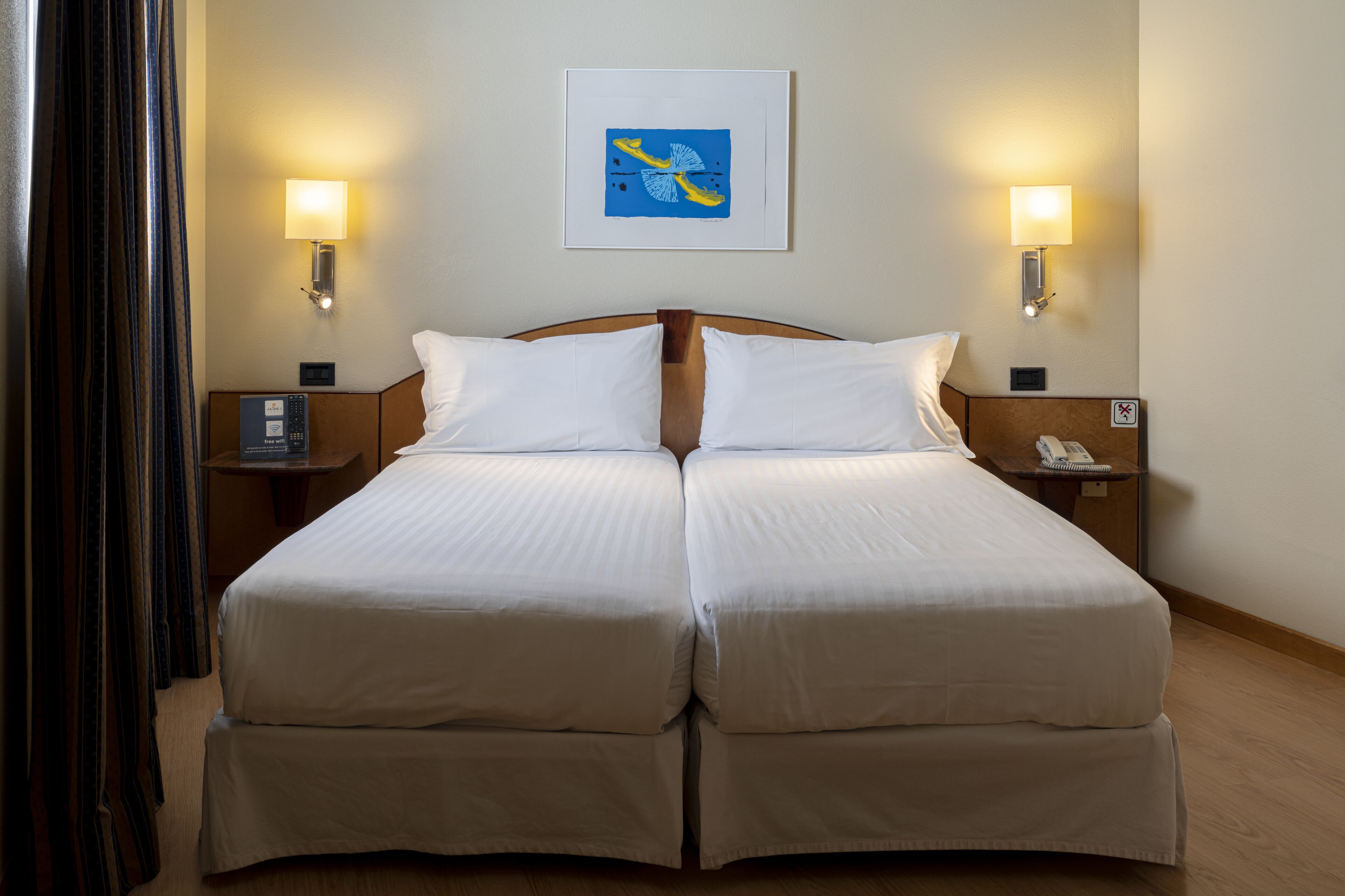 Cama doble con ropa de cama blanca, dos mesitas de noche con lámparas, una obra enmarcada sobre la cama y suelo de madera.