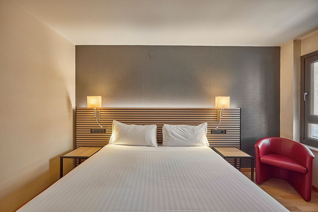 A hotel room with a large bed, two bedside tables, a red chair, and a wall-mounted light fixture.
