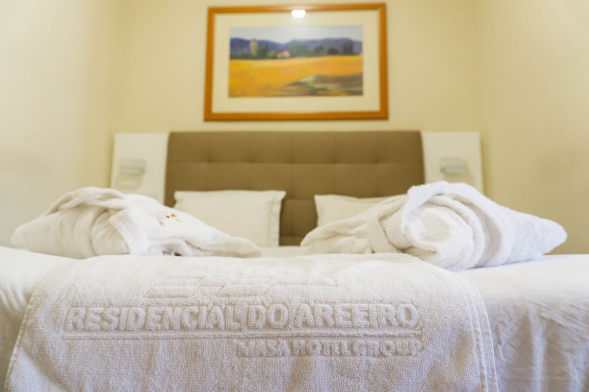 A neatly made bed with a white bedspread and two rolled towels. A framed painting hangs above the bed.