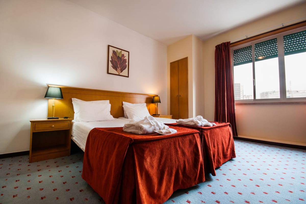 A hotel room with a double bed, two bedside tables, a nightstand, a window with curtains, and a wall-mounted artwork.