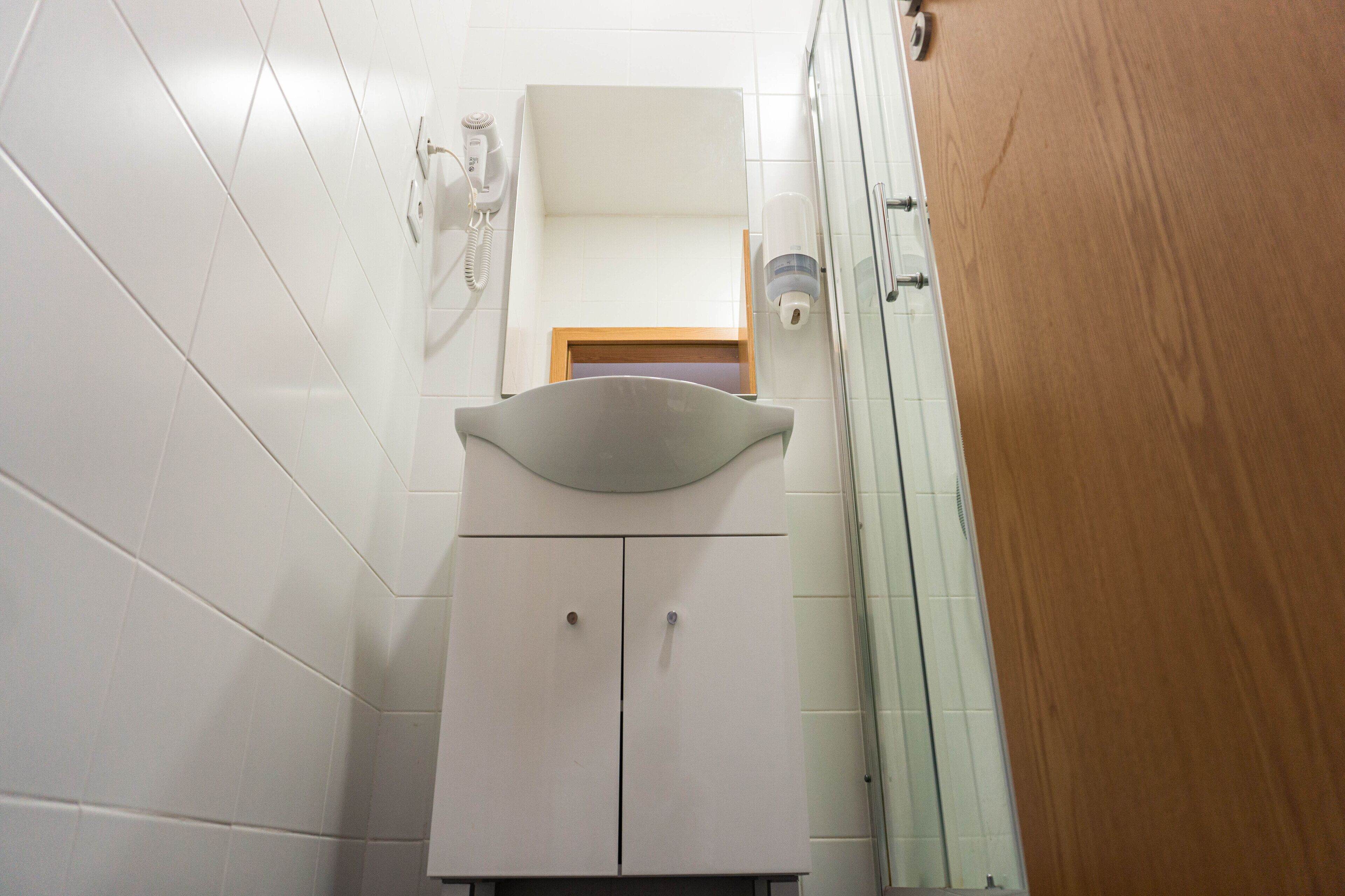 Un baño con paredes de azulejos blancos, un lavabo blanco con encimera curva y una puerta de madera.