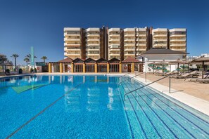 A swimming pool with clear blue water, surrounded by a building with multiple balconies and a covered area with umbrellas. - Occidental Fuengirola (Fuengirola)