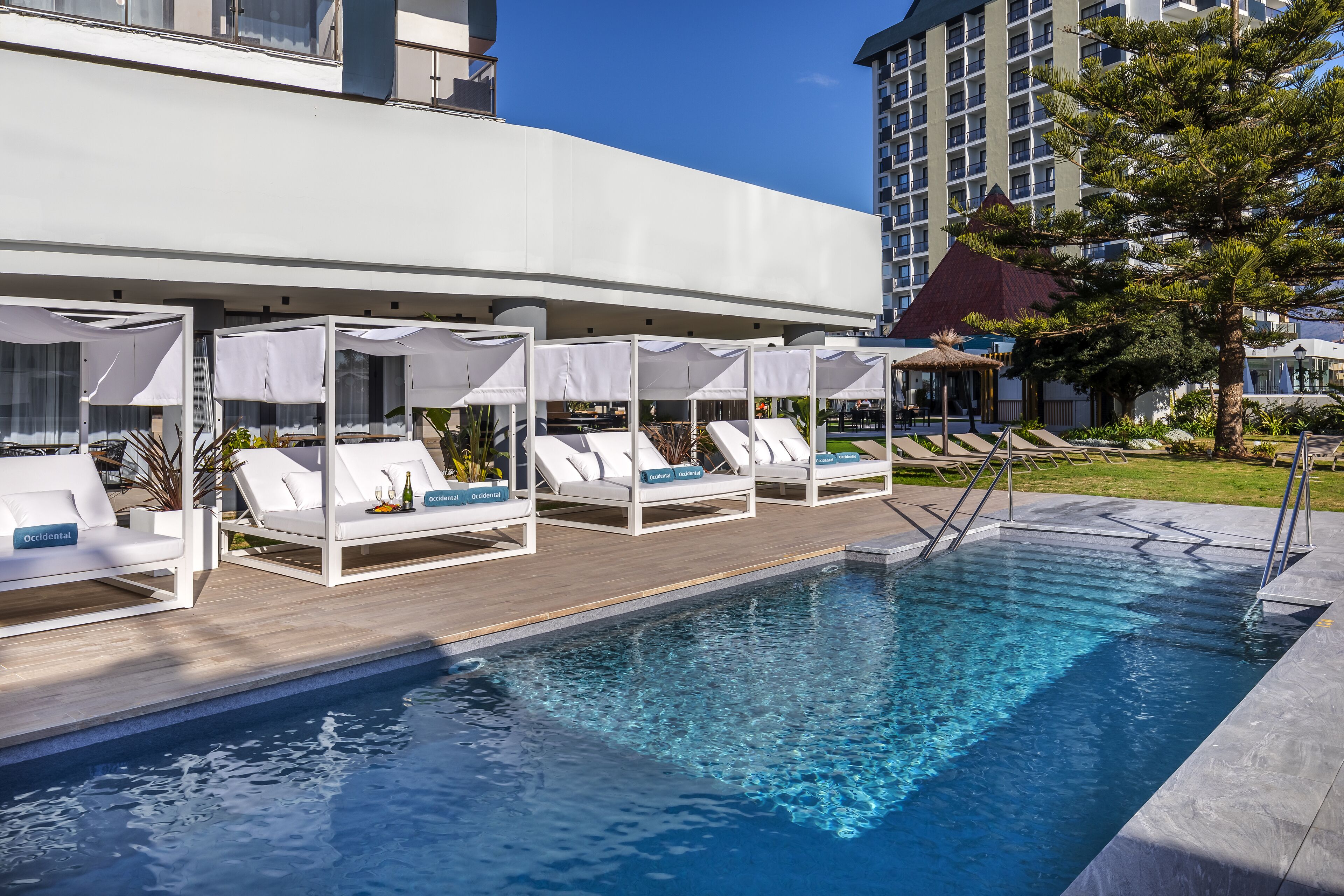 A poolside area with lounge chairs and umbrellas, a clear pool, and a building in the background.